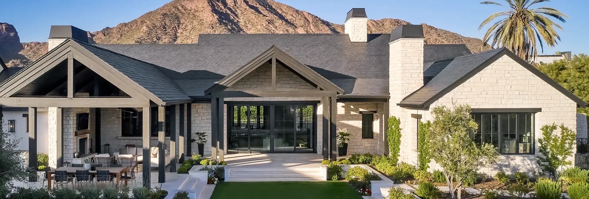 Luxury modern farmhouse with stone exterior, dark gabled roof, covered patio and outdoor dining overlooking landscaped yard and mountain backdrop