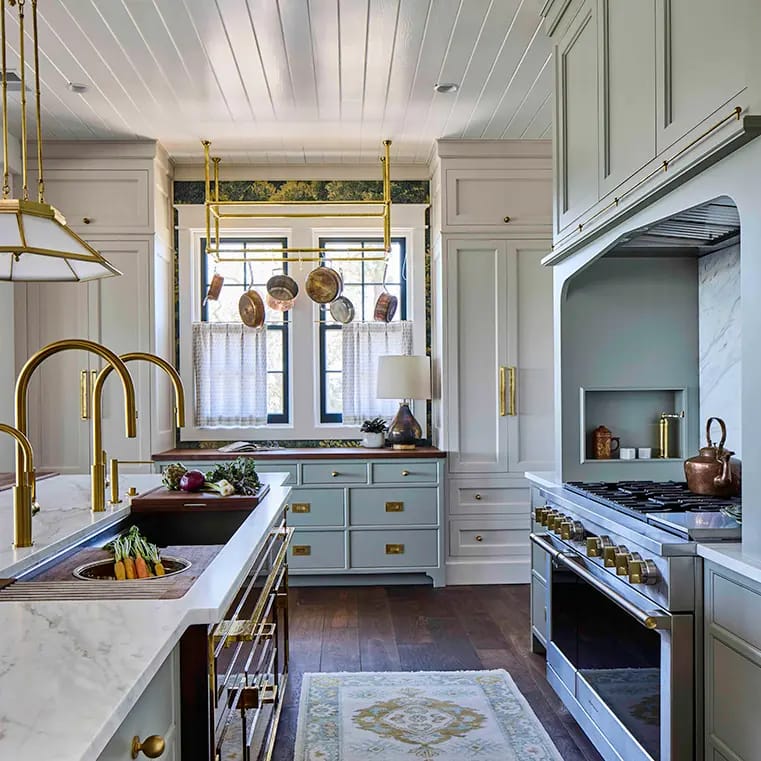 Bright luxury kitchen with light-blue cabinetry, marble countertops, brass hardware, island farmhouse sink, hanging copper pots, stainless gas range, dark hardwood floors.