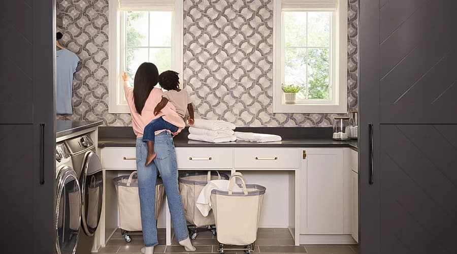 A woman holding a child in a quality laundry room with patterned wallpaper and modern appliances.