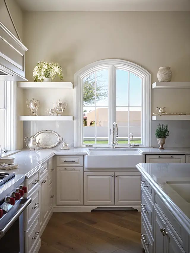 Bright white classic kitchen with farmhouse sink beneath arched window, marble countertops, open shelves displaying silver serving pieces and decorative accents.