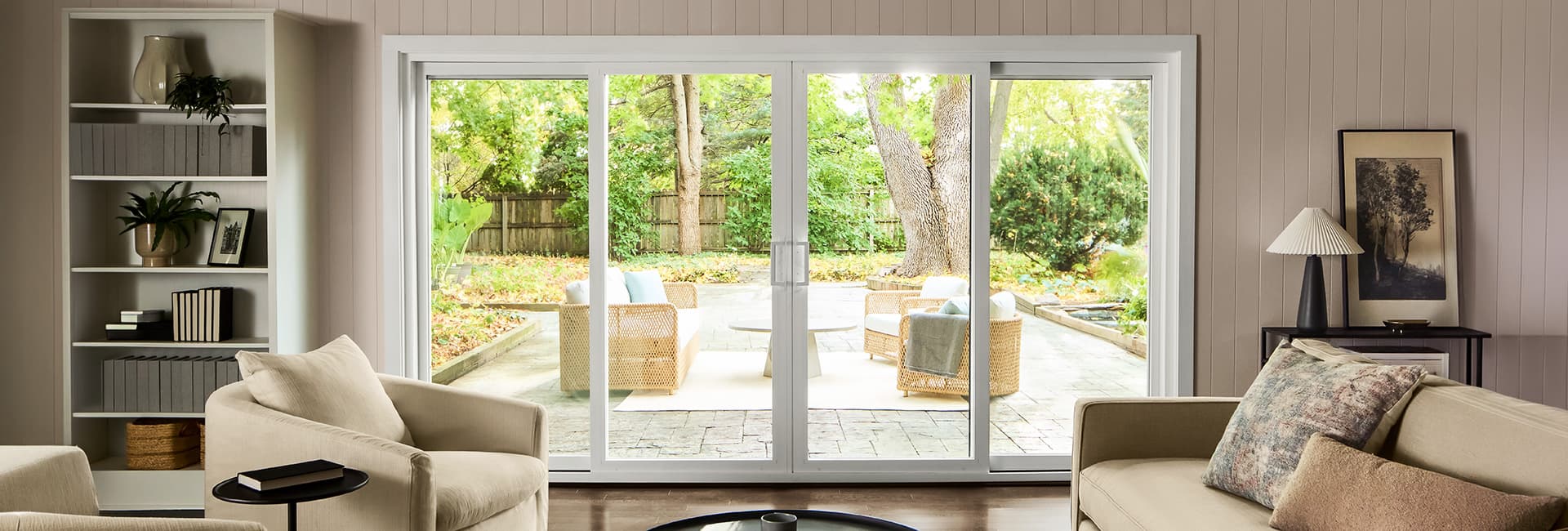 Living room with beige sofas and armchairs, sliding glass doors opening to a backyard patio with wicker chairs and trees.