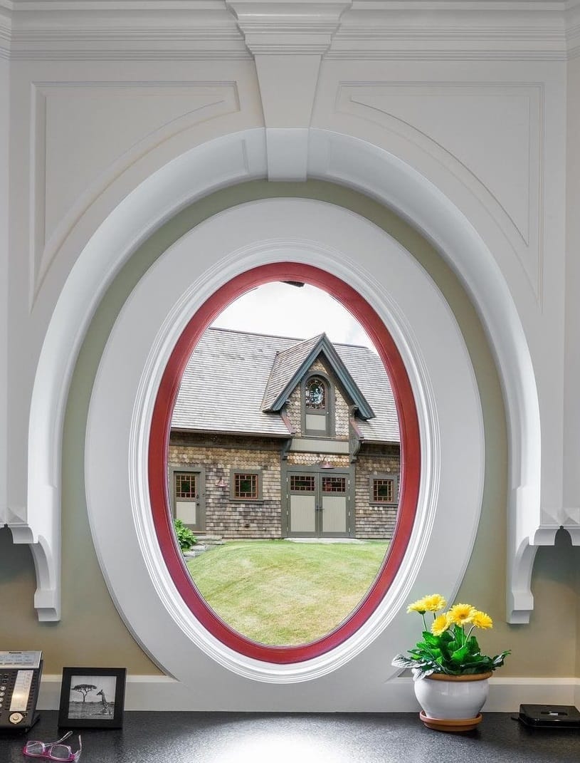 A red oval window with white trim sits over a desk and overlooks a house across the yard.