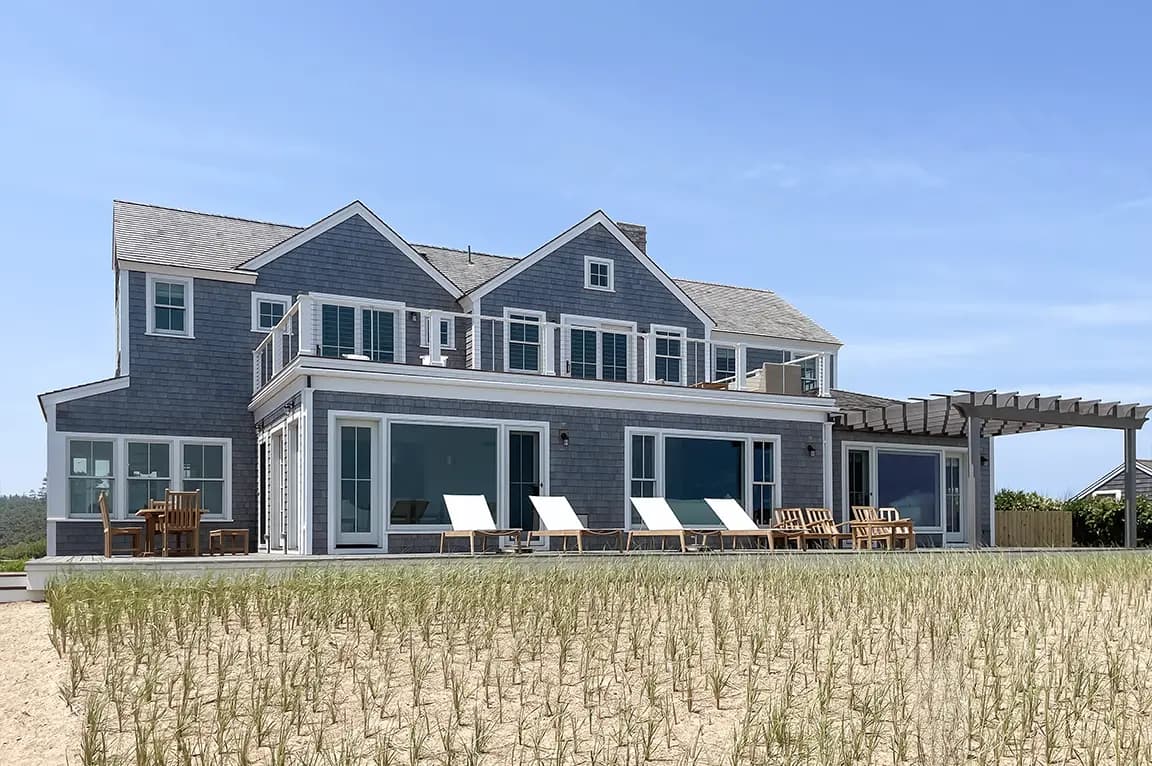 Large coastal shingle-style beachfront house with sliding glass doors, sun loungers on the deck, pergola, and sandy dune grasses under a blue sky.