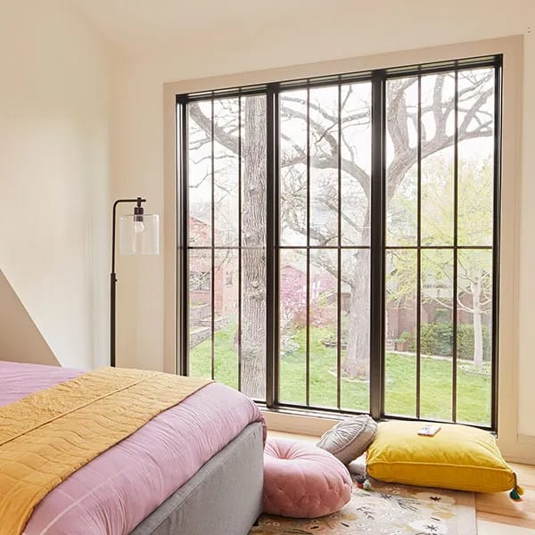 A bedroom with a large black-framed window and colorful pillow and blanket combinations on the bed and floor.