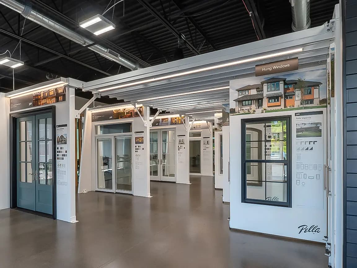 Pella window showroom interior with multiple hung window and door displays on white panels under exposed industrial ceiling lighting
