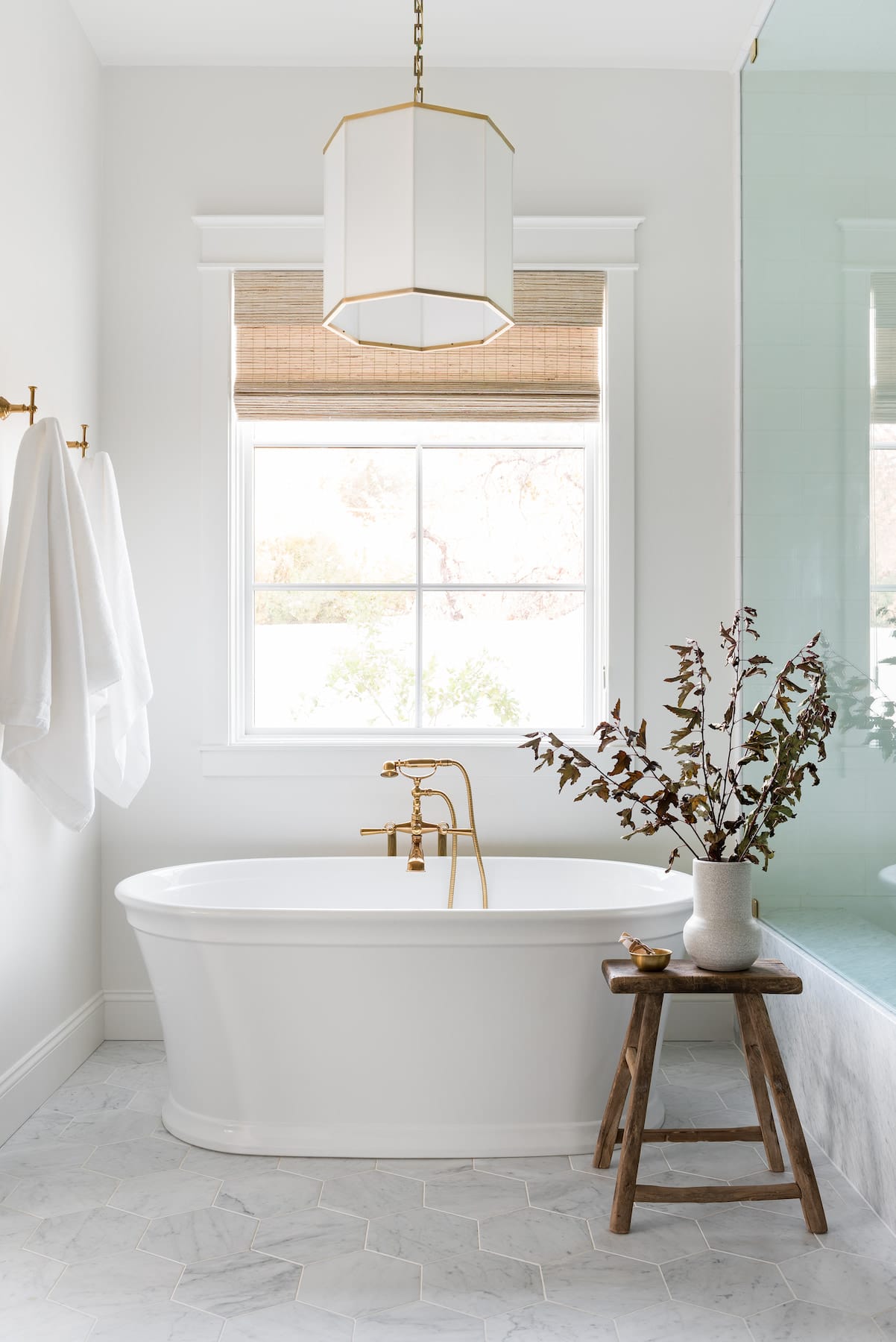 A white picture window with Roman shades sits behind a freestanding bathtub.
