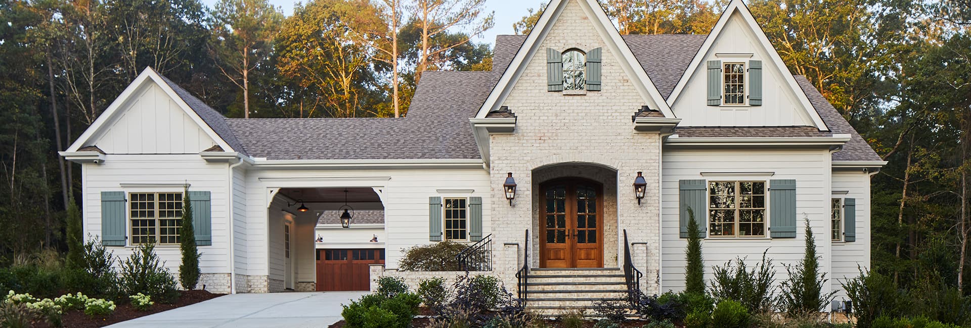 Modern white farmhouse with gabled roof, brick entryway and wooden double doors, blue shutters, attached garage and landscaped yard.