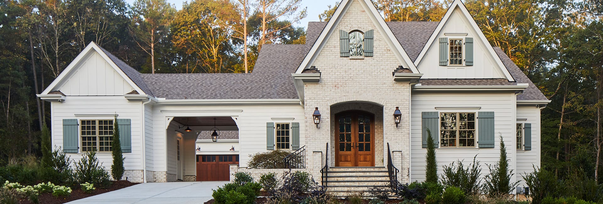 Modern white farmhouse with gabled roof, brick entryway and wooden double doors, blue shutters, attached garage and landscaped yard.