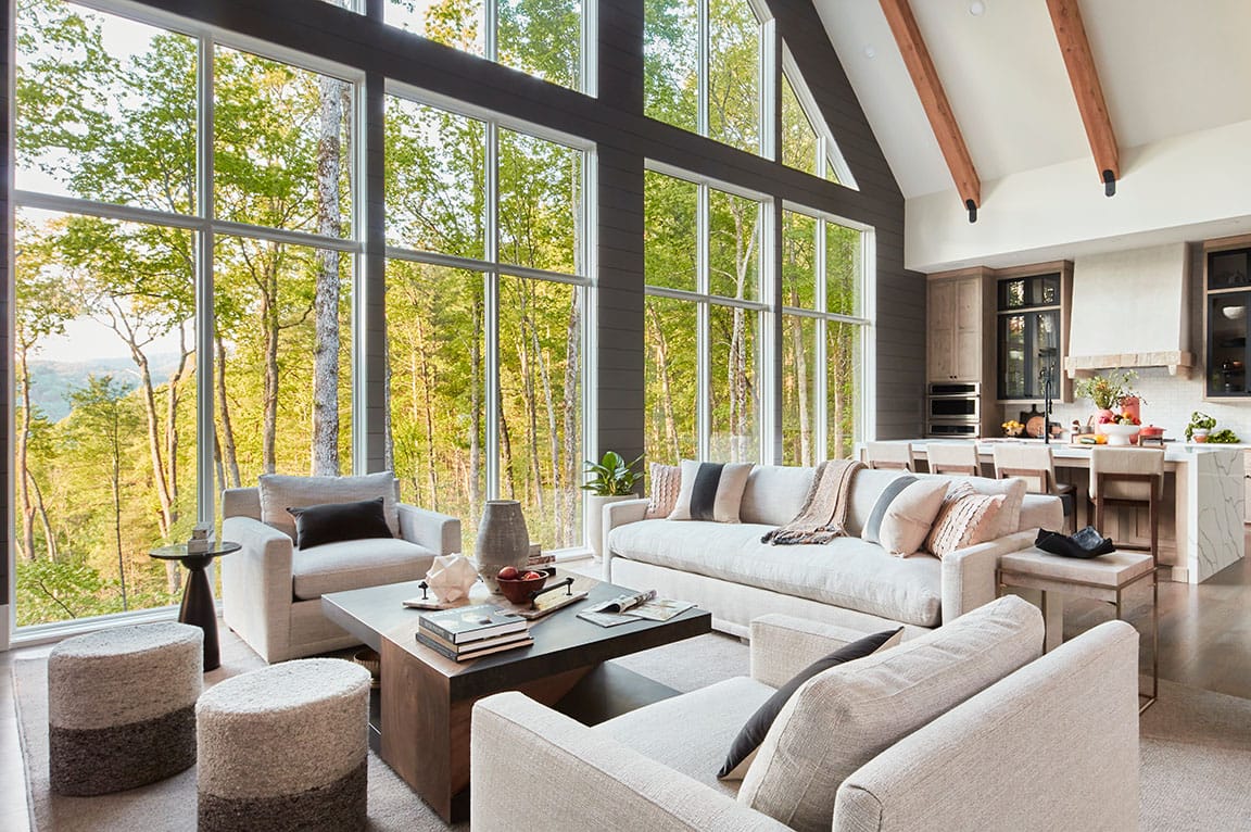 Modern living room with floor-to-ceiling windows, neutral couches, and forest views.