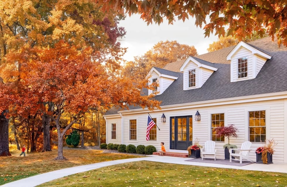White suburban house with dormer windows and American flag, front porch with chairs and walkway, surrounded by vibrant orange autumn foliage