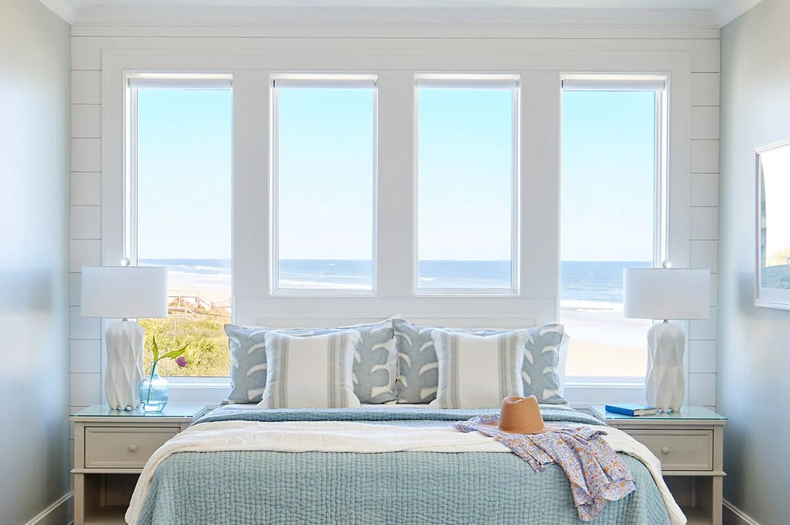 A bright coastal bedroom with large windows overlooking the ocean, featuring blue bedding and beach decor, no hurricane visible.