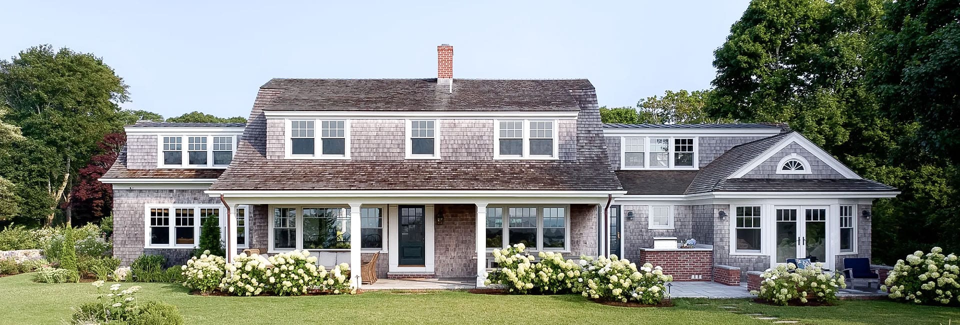 Two-story cedar shingle-style coastal house with a covered front porch, white trim, hydrangea borders and manicured lawn