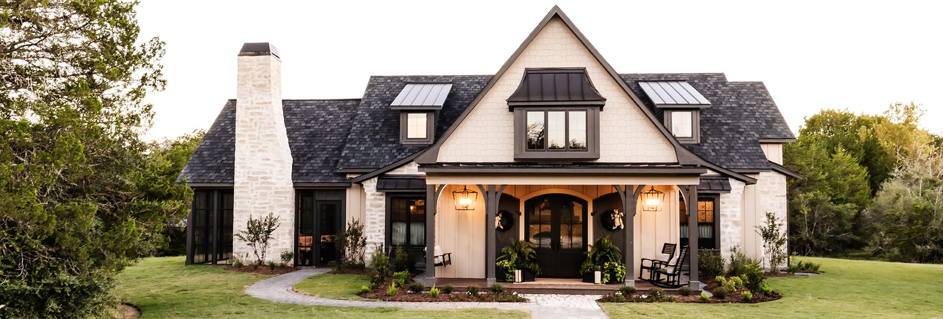 Modern farmhouse exterior with gabled roof and stone chimney, covered front porch with lanterns, rocking chairs, and landscaped lawn