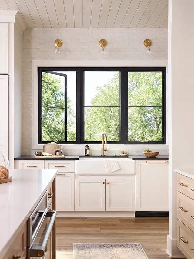 Bright modern farmhouse kitchen with black-framed windows, white cabinets, apron-front sink, brass faucet and light wood floors.