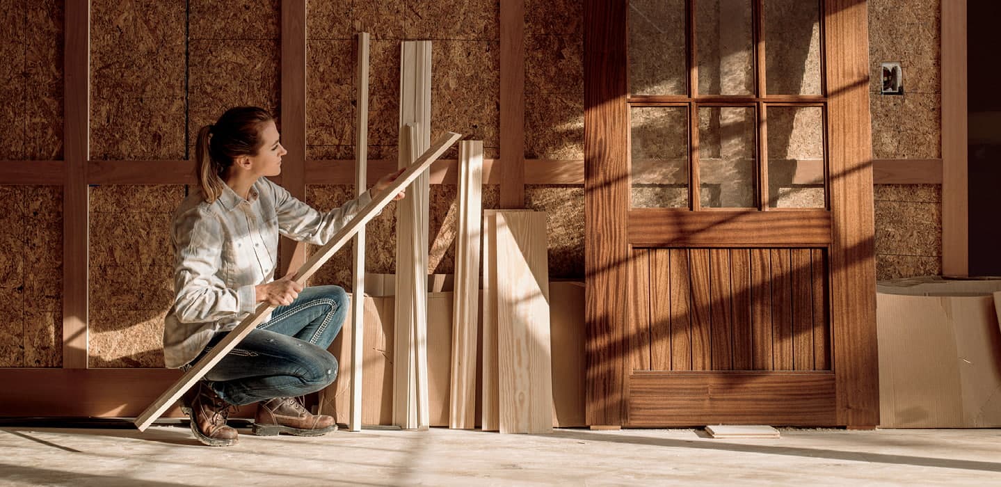 a female construction work is examining a piece of trim by a wood entry door
