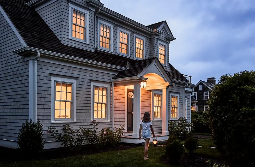 Two-story shingle-style house at dusk with warm-lit windows and a person carrying a lantern walking toward the front porch.