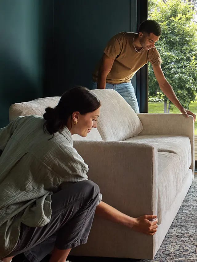 Two people moving a beige sofa into a modern living room by a large glass door overlooking a green yard.