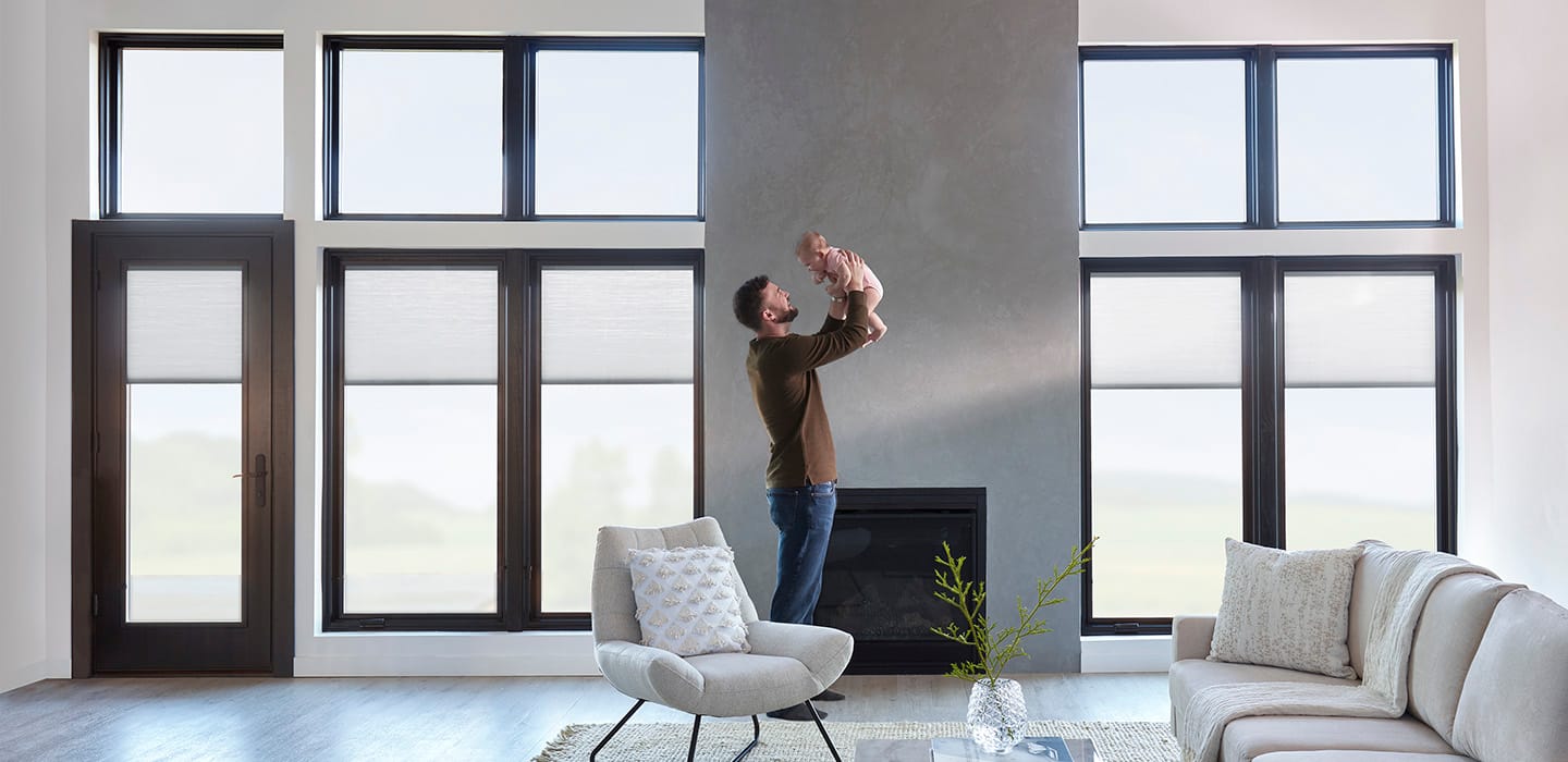 A father is playing with his baby daughter in a minimalist living room with large black windows on the wall behind them.