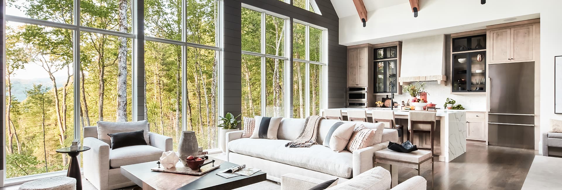 Open-concept living room and kitchen with floor-to-ceiling windows overlooking a forest, neutral sofas and marble island