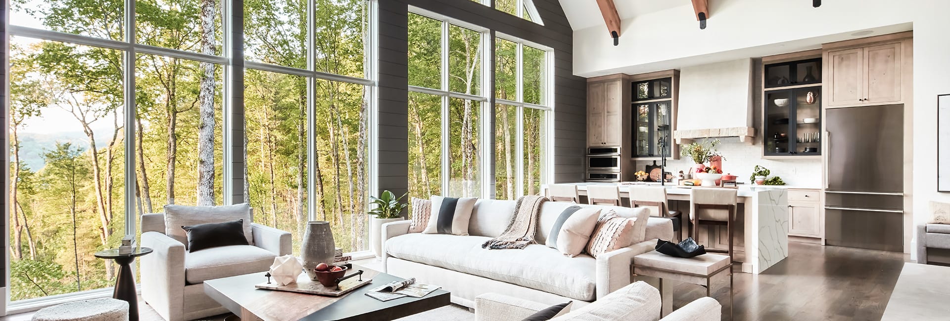 Open-concept living room and kitchen with floor-to-ceiling windows overlooking a forest, neutral sofas and marble island