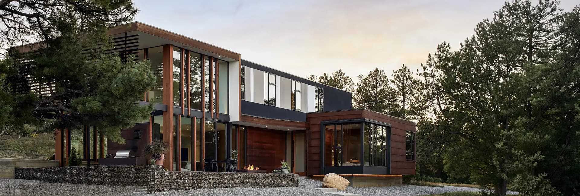 Modern glass-and-wood house with floor-to-ceiling windows, outdoor patio and fire pit, nestled in a pine forest at dusk