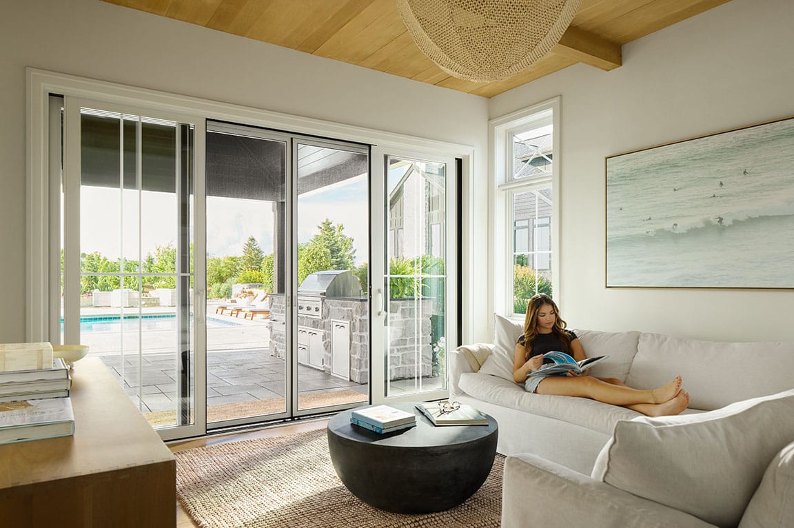 A woman is sitting on a sofa next to a white four-panel sliding patio door.