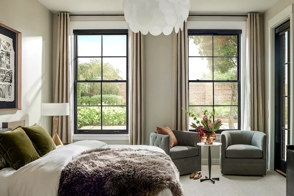Modern neutral bedroom with large black-framed windows and garden view, bed with faux-fur throw and green velvet pillows, two gray armchairs and side table with vase of flowers.