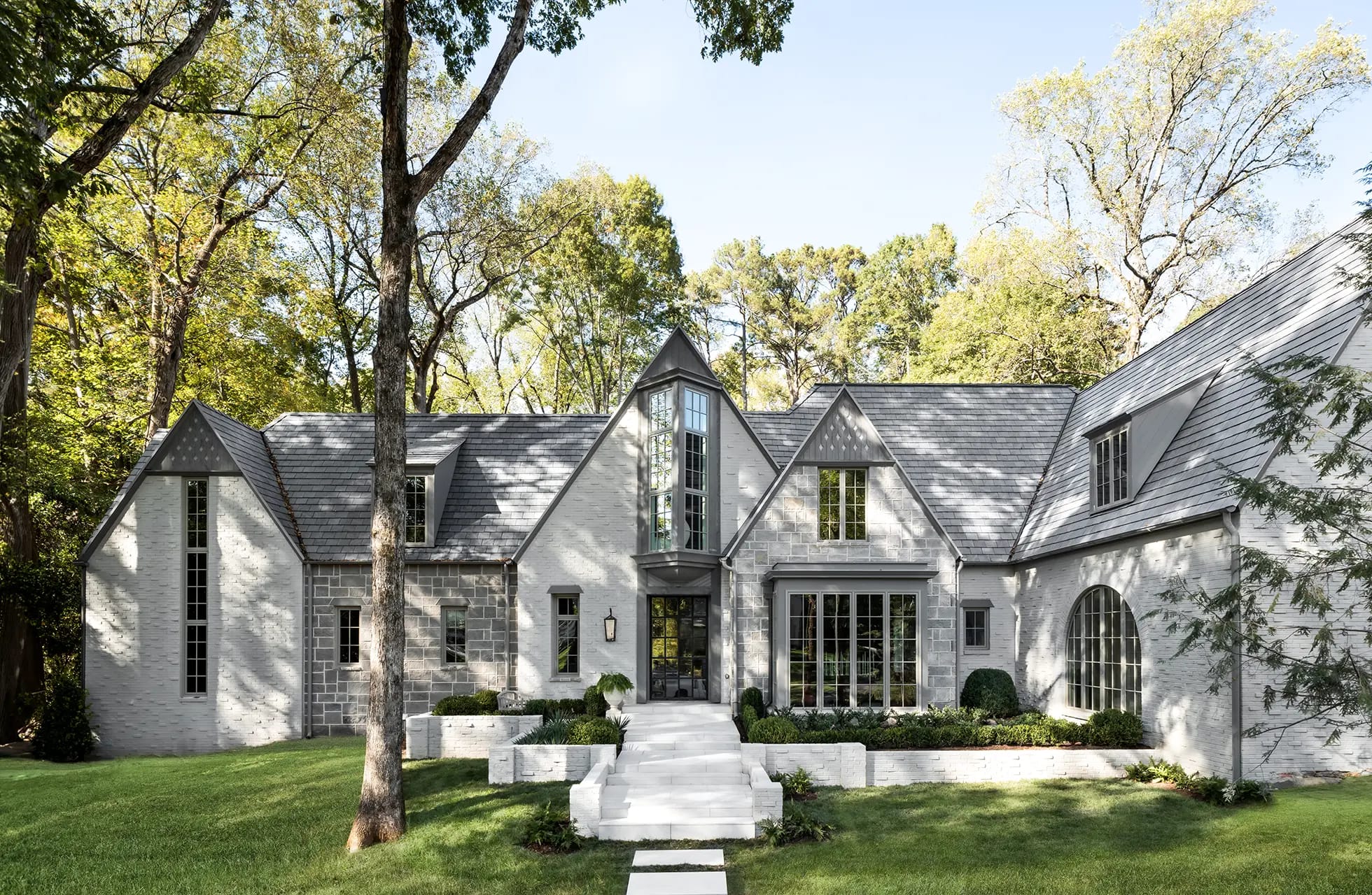 Luxury white brick Tudor-style house with slate roof, tall central multi-pane window tower, landscaped front steps and wooded lawn