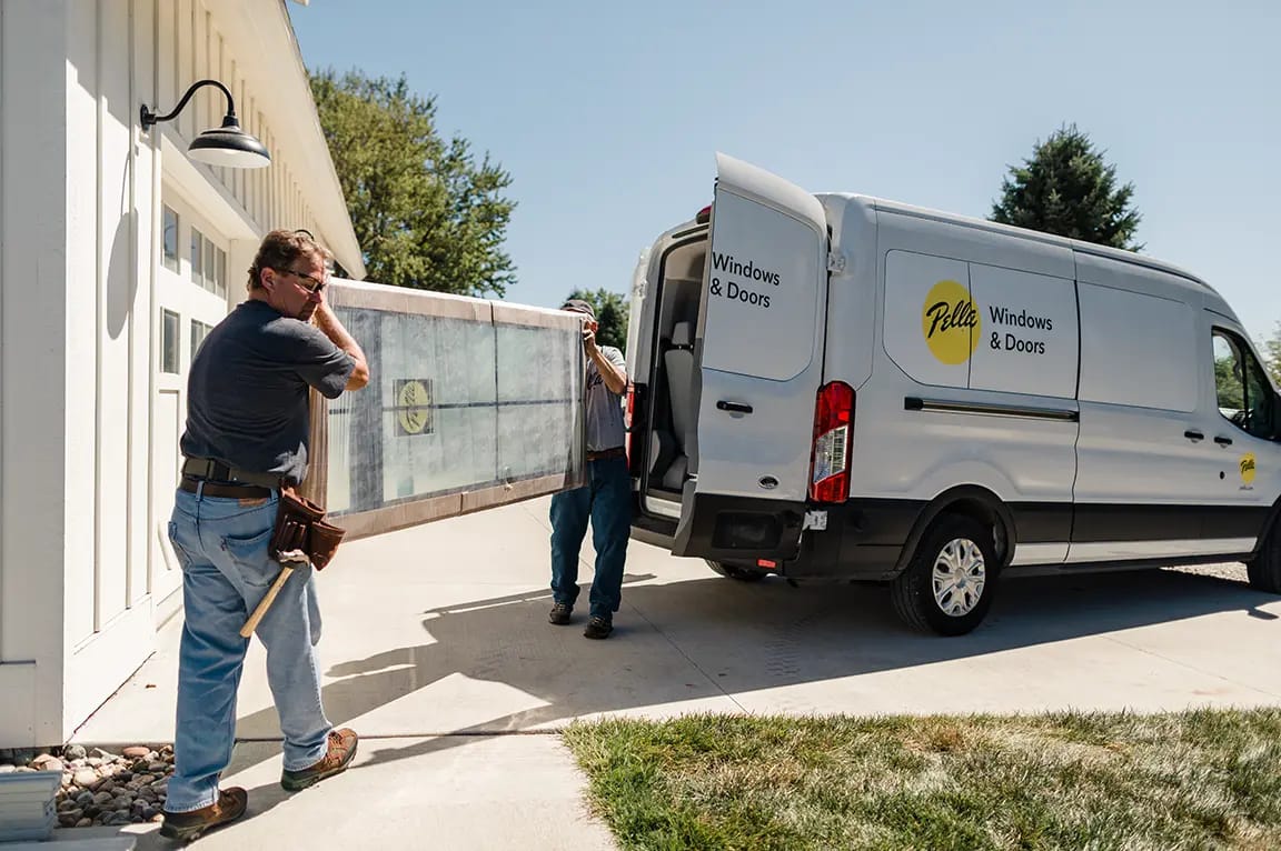 Pella windows installers carrying a large window panel from a branded service van to a suburban home's garage driveway.