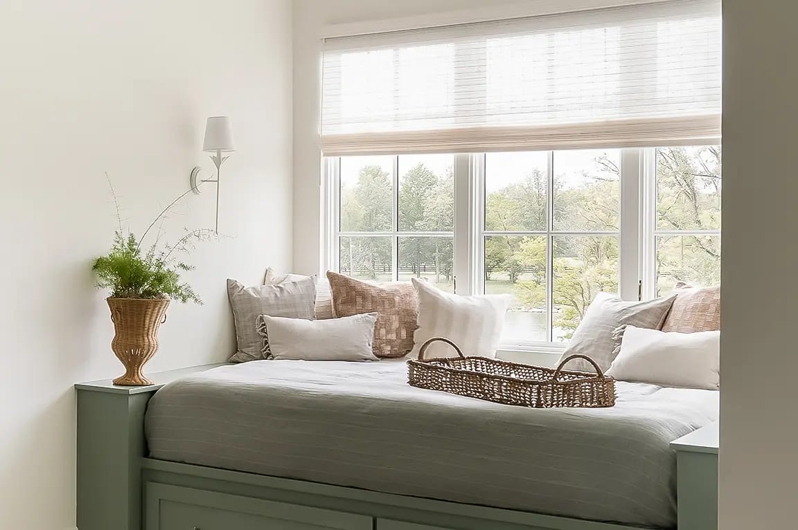 Sunlit built-in window seat daybed with neutral pillows, wicker tray and vase of greenery overlooking a tree-lined view.