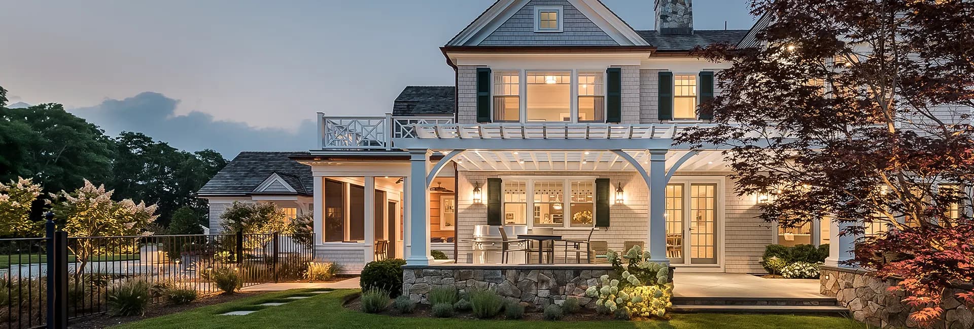 Shingle-style luxury home exterior at dusk with illuminated covered patio and white pergola, outdoor dining area, stone foundation and landscaped yard