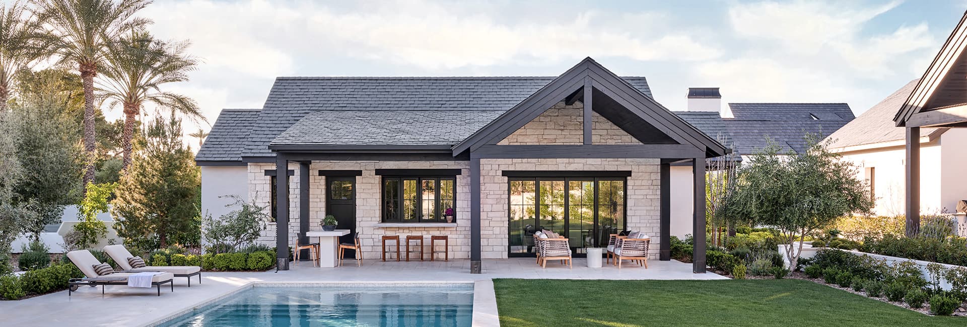 Modern stone house backyard with swimming pool, covered patio with outdoor seating and lounge chairs, landscaped yard and palm trees