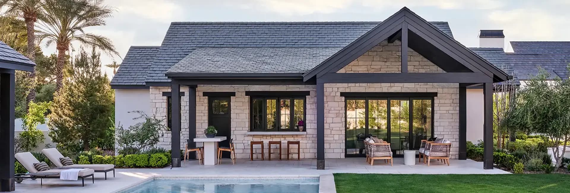 Modern stone house backyard with swimming pool, covered patio with outdoor seating and lounge chairs, landscaped yard and palm trees