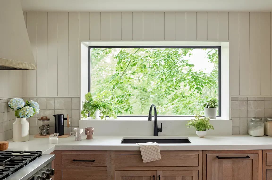 Kitchen sink with black faucet under large picture window overlooking green trees, white countertop, wood cabinets and potted plants.