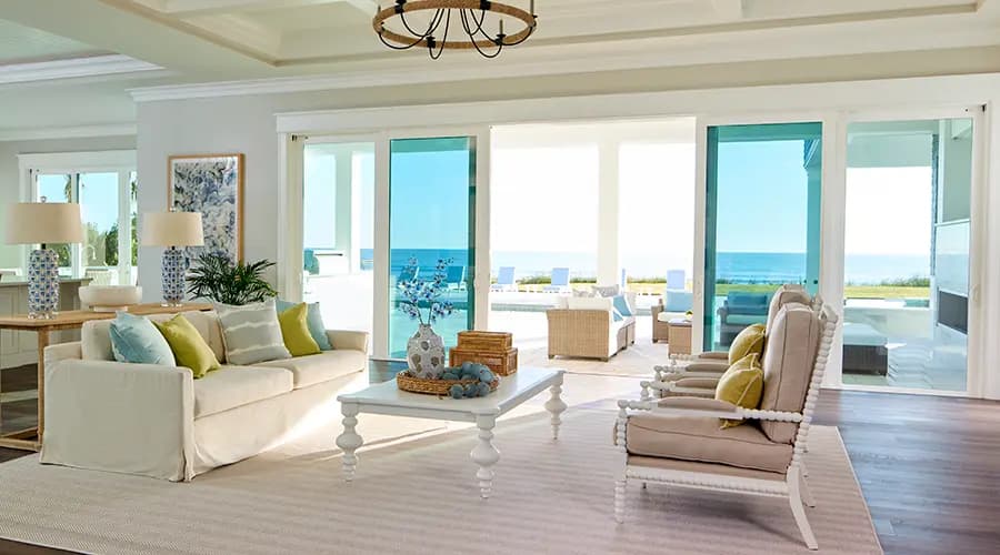Coastal living room in a beach house with beige sofa, pastel accent pillows, white coffee table and ocean view through sliding glass doors.