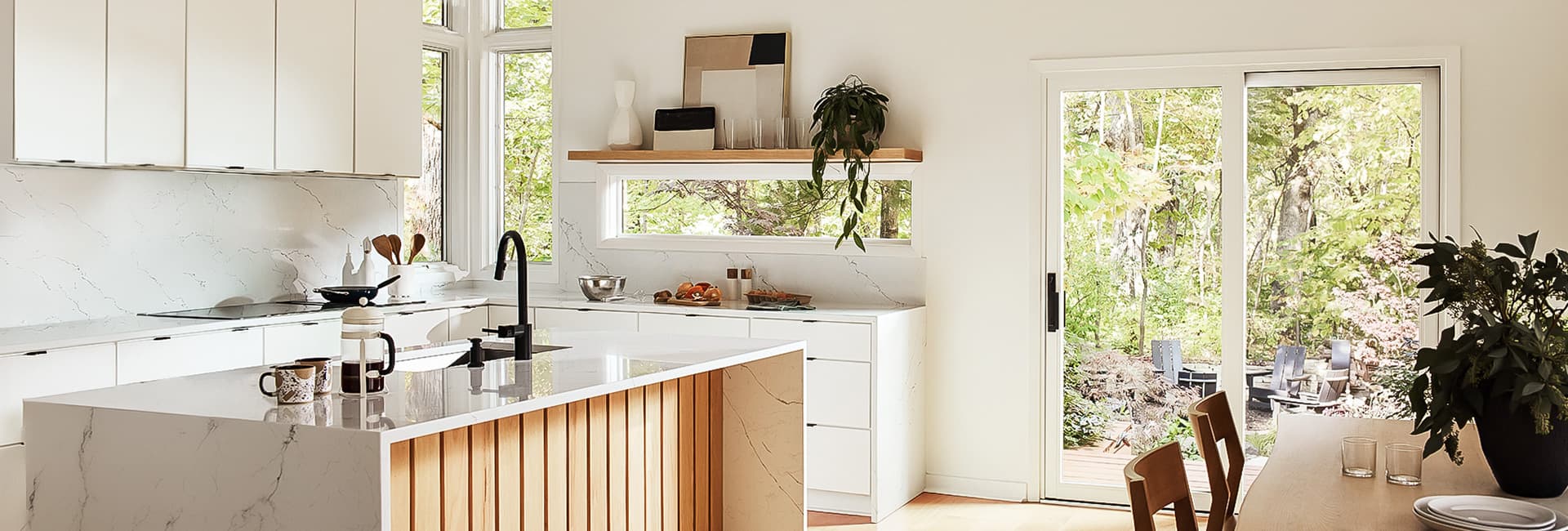 Modern white kitchen with marble countertops, wood-slat island and black faucet, sliding glass door to wooded backyard patio.