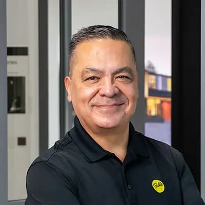 Smiling man in black polo shirt with yellow logo, arms crossed in a modern window showroom