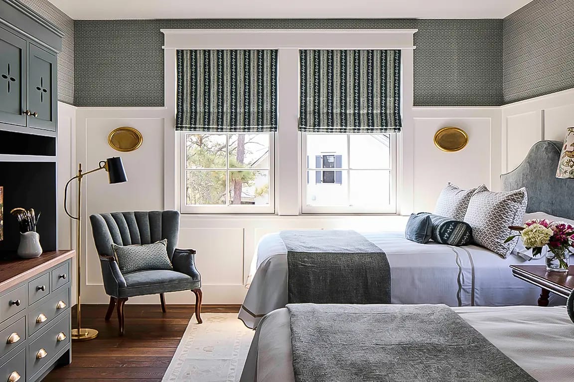 Gray-and-white guest bedroom with two beds, dark gray throws, green patterned Roman shades, velvet accent chair and brass floor lamp.