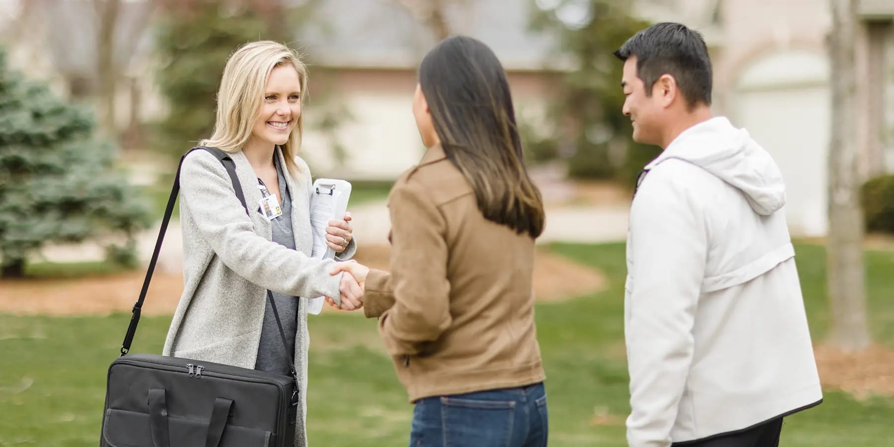 Field representative with clipboard and shoulder bag shaking hands with a woman outside a suburban home while a man stands nearby.