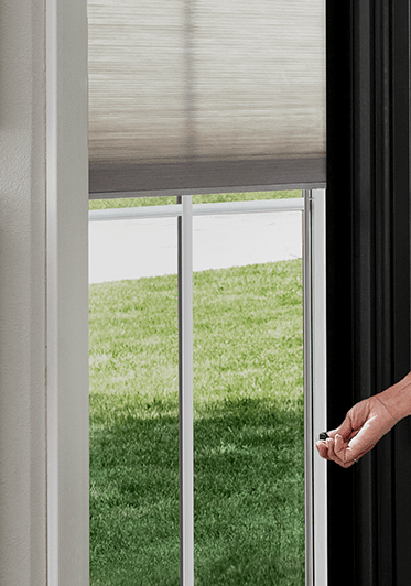 A woman's hand is raising between-the-glass white blinds to show the green grass outside.