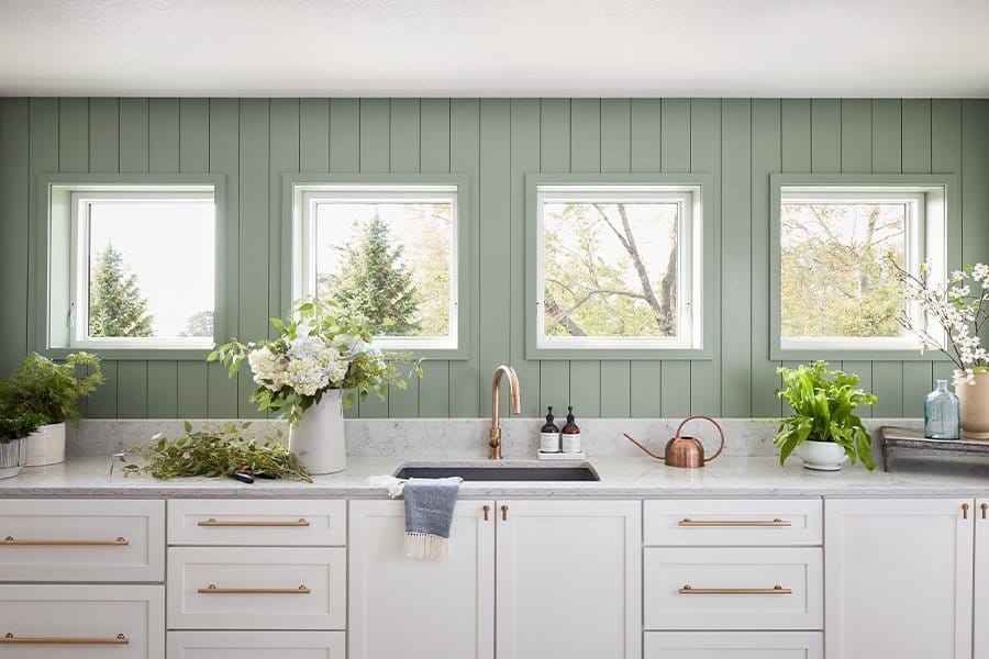 Modern farmhouse kitchen with white cabinets, marble countertop, brass faucet, green shiplap wall and four square windows.