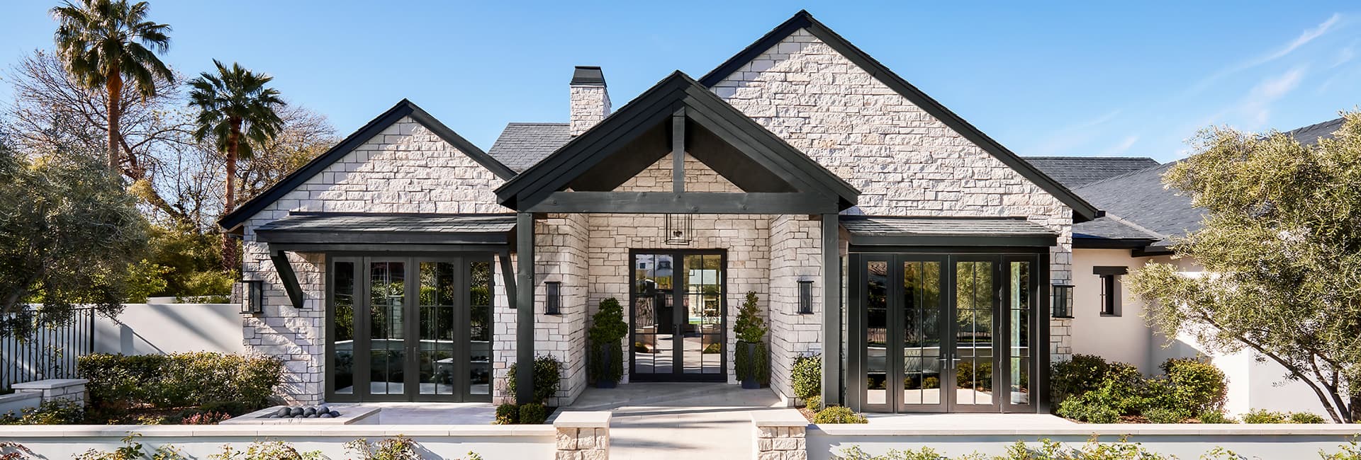 Modern stone house exterior with black-trimmed gabled entrance, large glass doors and landscaped front yard with palm trees.