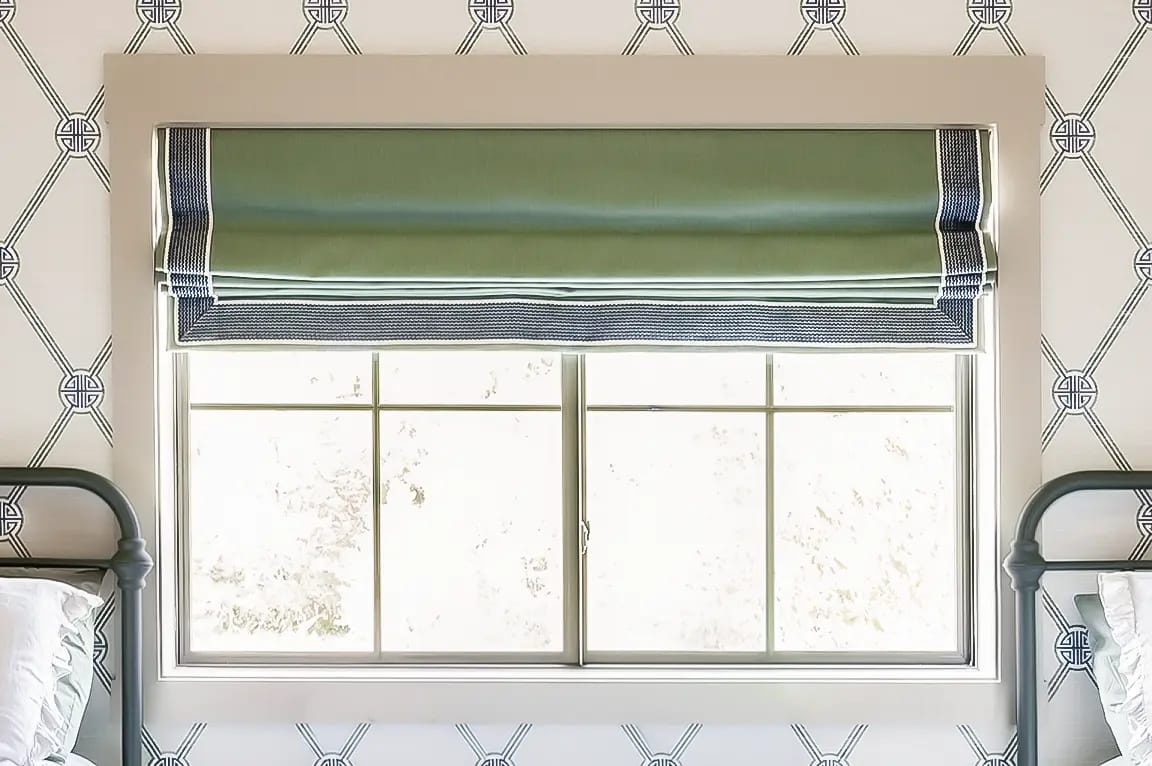 Bedroom window with green Roman shade and navy trim, geometric wallpaper and twin metal bed frames.