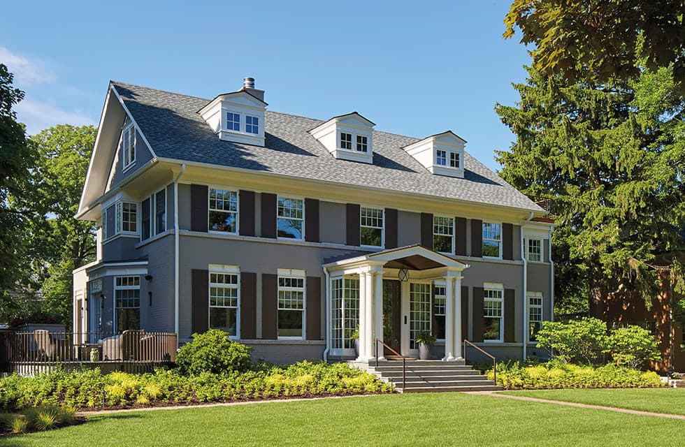 Two-story gray colonial-style house with dormer windows, columned front porch and landscaped lawn