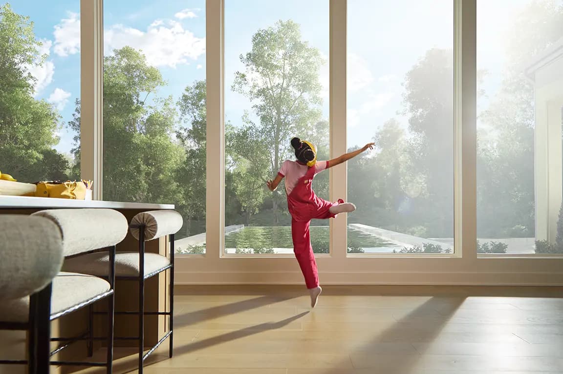 Little girl dancing on a sunlit hardwood floor in a modern living room with floor-to-ceiling windows overlooking a backyard pool and trees