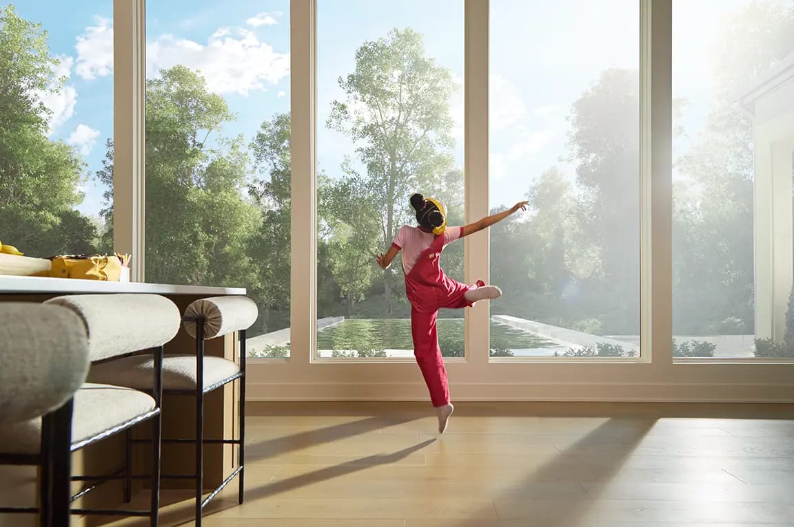 Little girl dancing on a sunlit hardwood floor in a modern living room with floor-to-ceiling windows overlooking a backyard pool and trees