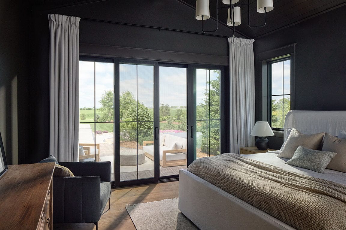 Modern bedroom with black-framed sliding glass patio doors opening to an outdoor seating area and landscape view.