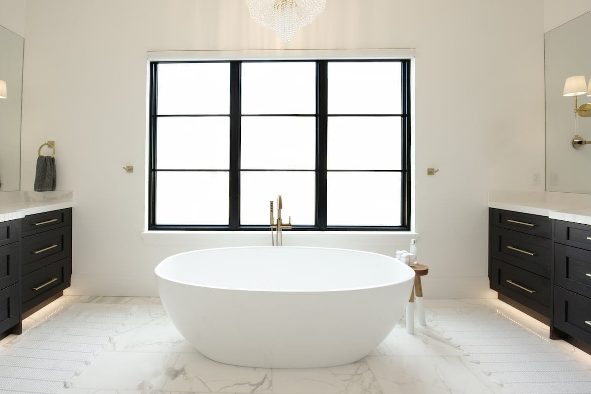 A black picture window sits over a white freestanding bathtub, with black vanities facing each other on opposite sides of the bathroom.