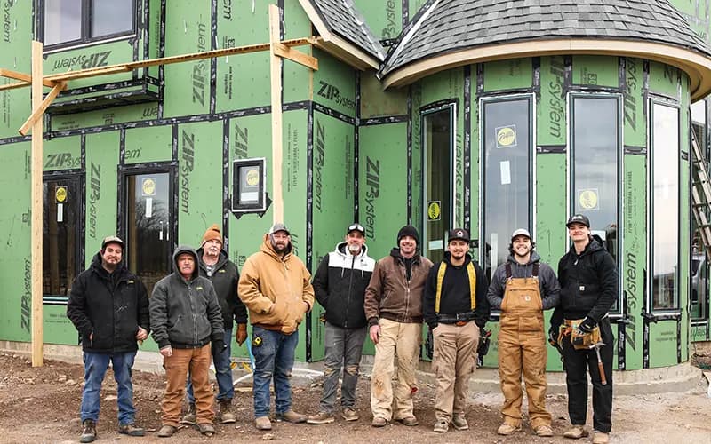 Construction crew standing in front of a new house wrapped in green ZIP System sheathing with installed tall windows and a rounded turret
