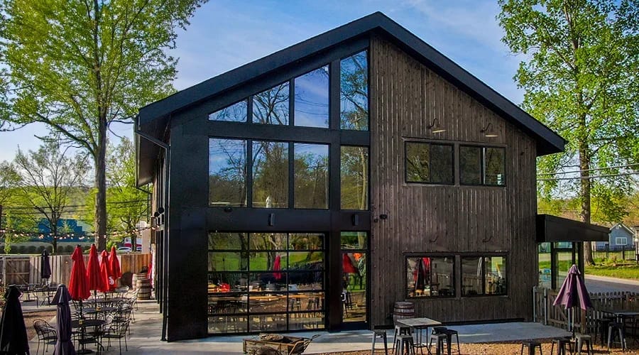 Contemporary building with large black-framed windows and dark wood siding, featuring outdoor seating and surrounded by trees.